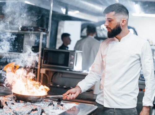 Frying Pan Is On Fire. Chef In White Uniform Cooking Food At Kitchen. Busy Day At Work